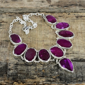 May include: A silver-toned necklace featuring large, oval-shaped, magenta-colored gemstones. The gemstones are set in a silver bezel and connected by a silver chain. The necklace is displayed on a wooden surface.