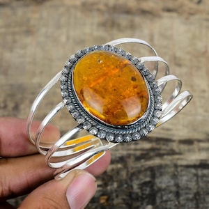May include: A silver cuff bracelet featuring a large, oval amber stone. The amber is a warm, golden color with visible inclusions. The stone is set in a detailed, ornate silver frame with a textured border. The bracelet has a simple, open design.