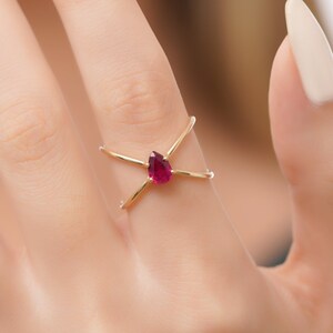 May include: A gold ring with a red teardrop-shaped gemstone. The ring has a criss-cross design.