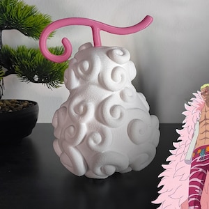 Peut inclure: Un modèle imprimé en 3D blanc du fruit du démon "Hito Hito no Mi, Modèle : Nika" de la série d'anime One Piece. Le fruit a une tige rose et est en forme de poire avec une surface texturée. Le fruit est à côté d'une image de dessin animé d'un personnage avec des cheveux roses et un manteau de fourrure rose.