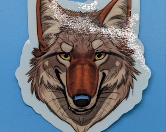 May include: A die-cut sticker featuring a stylized illustration of a wolf head. The wolf has brown and gray fur, pointed ears with reddish-brown inner details, and piercing yellow eyes. The nose is dark brown with a small blue highlight.