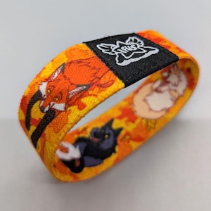 Puede incluir: Una pulsera tejida amarilla, naranja y negra con un diseño de zorro, gato y perro blanco. La pulsera tiene una etiqueta negra con la palabra "HUG" en letras blancas.