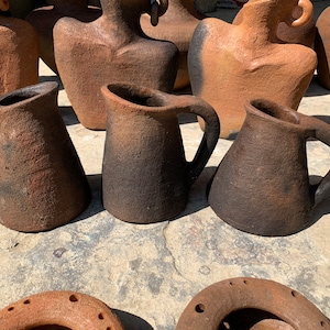 Pode incluir: Uma coleção de jarras e vasos de cerâmica castanha rústica feitos à mão. As jarras têm uma pega e um bico, enquanto os vasos têm uma forma arredondada. A cerâmica tem uma superfície texturizada e um gradiente de castanho escuro a castanho claro.