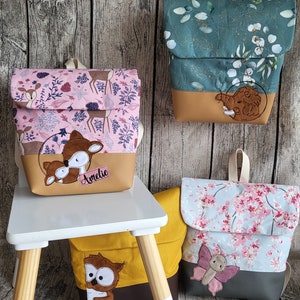 Könnte beinhalten: Vier Rucksäcke mit Stoffklappen mit Mustern aus Hirschen, Blättern und Blumen. Die Taschen haben einfarbige Unterteile in Beige, Braun oder Grau. Eine Tasche zeigt eine braune Hirsch-Applikation mit dem Namen Amelie, eine andere ein Mammut und eine eine Eule.