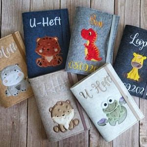 Könnte beinhalten: Eine Auswahl an Filz-Buchhüllen in verschiedenen Farben, jede mit einem gestickten Tiermotiv. Die Hüllen zeigen einen Tiger, einen Dinosaurier, eine Giraffe, ein Nilpferd, einen Bären und einen Frosch. Jede Hülle hat einen Namen oder Text aufgestickt.