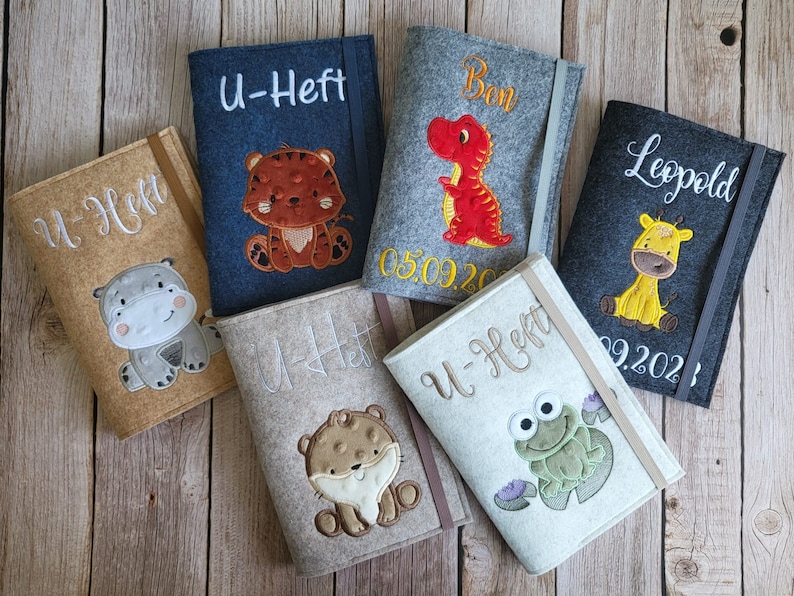 K&ouml;nnte beinhalten: Eine Auswahl an Filz-Buchh&uuml;llen in verschiedenen Farben, jede mit einem gestickten Tiermotiv. Die H&uuml;llen zeigen einen Tiger, einen Dinosaurier, eine Giraffe, ein Nilpferd, einen B&auml;ren und einen Frosch. Jede H&uuml;lle hat einen Namen oder Text aufgestickt.