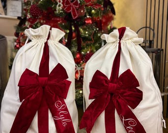 Pode incluir: Dois sacos de presente de tecido na cor creme com fecho de cordão, cada um com um grande laço de veludo vermelho escuro e uma faixa vertical de veludo vermelho. O nome Lyla está bordado num laço e o nome Leo no outro.