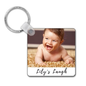 May include: Square keyring with a photograph of a smiling baby. The baby is lying on a textured surface and has brown hair. The text "Lily's Laugh" is written in cursive at the bottom of the keyring.