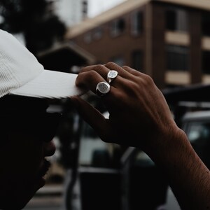 May include: A person wearing a white hat and two silver rings with intricate designs on their fingers.