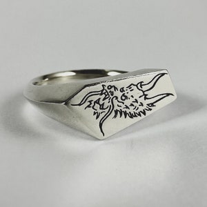 May include: A silver ring featuring a dragon head design engraved on the top. The ring is a signet style with a flat top and a rounded band.