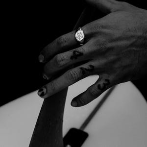 May include: A silver signet ring with a floral design on the face. The ring is worn on the index finger of a hand.