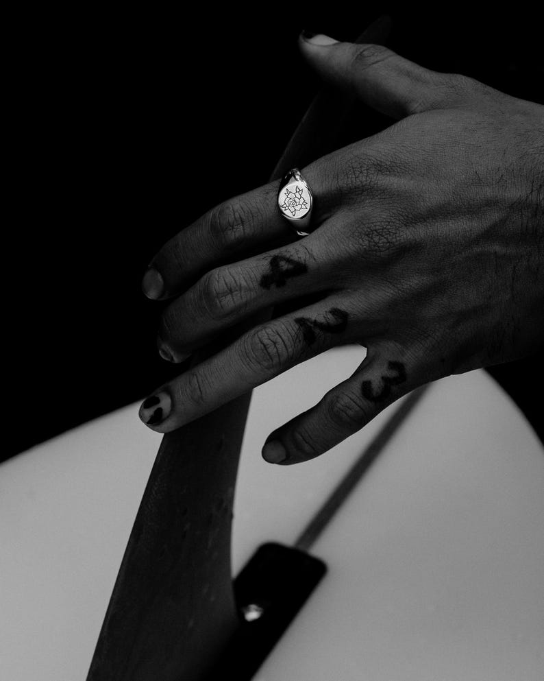 May include: A silver signet ring with a floral design on the face. The ring is worn on the index finger of a hand.