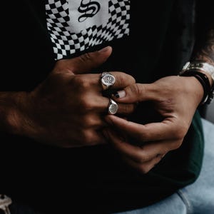 May include: Two silver signet rings with detailed designs on the face of the rings. The rings are worn on the index and middle fingers of a person's hand.