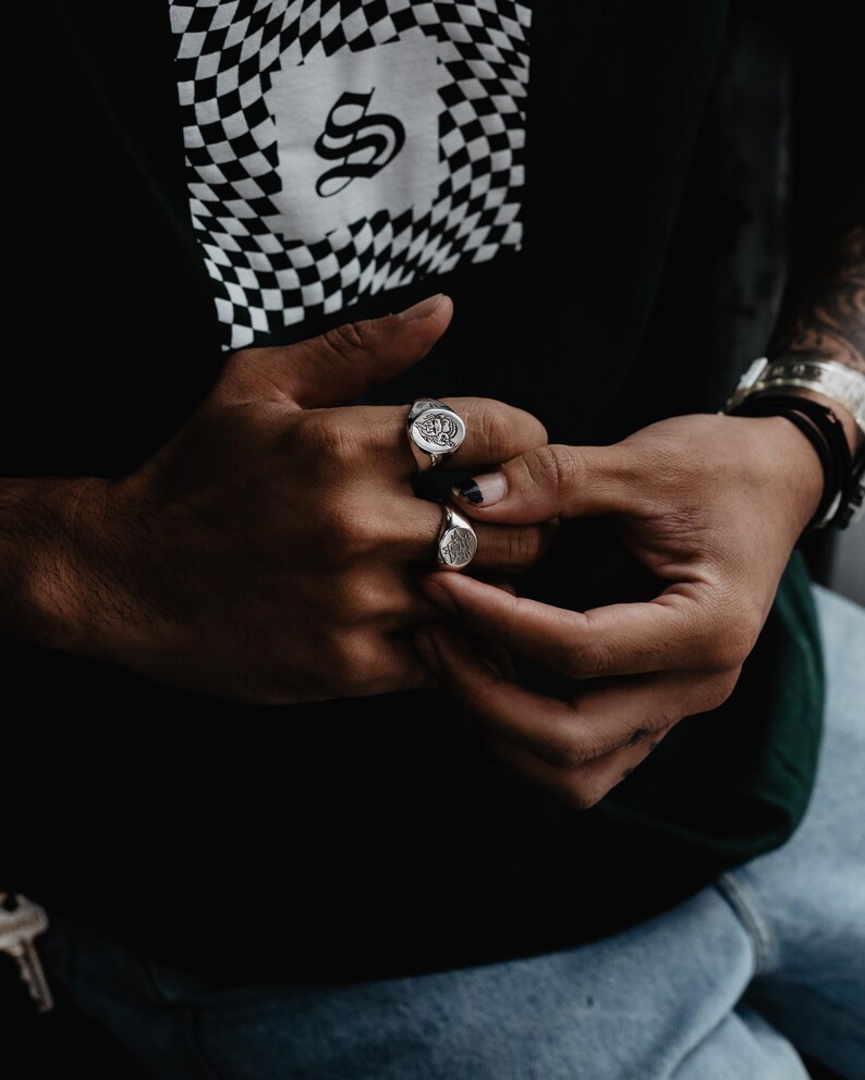 May include: Two silver signet rings with detailed designs on the face of the rings. The rings are worn on the index and middle fingers of a person's hand.