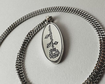 Op de afbeelding: Een zilveren ketting met een zilveren ovale hanger met een zwart-witte lijntekening van een roos.