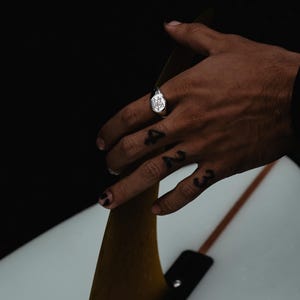 May include: A silver signet ring with a detailed design on the face. The ring is worn on the index finger of a hand resting on a surfboard fin.