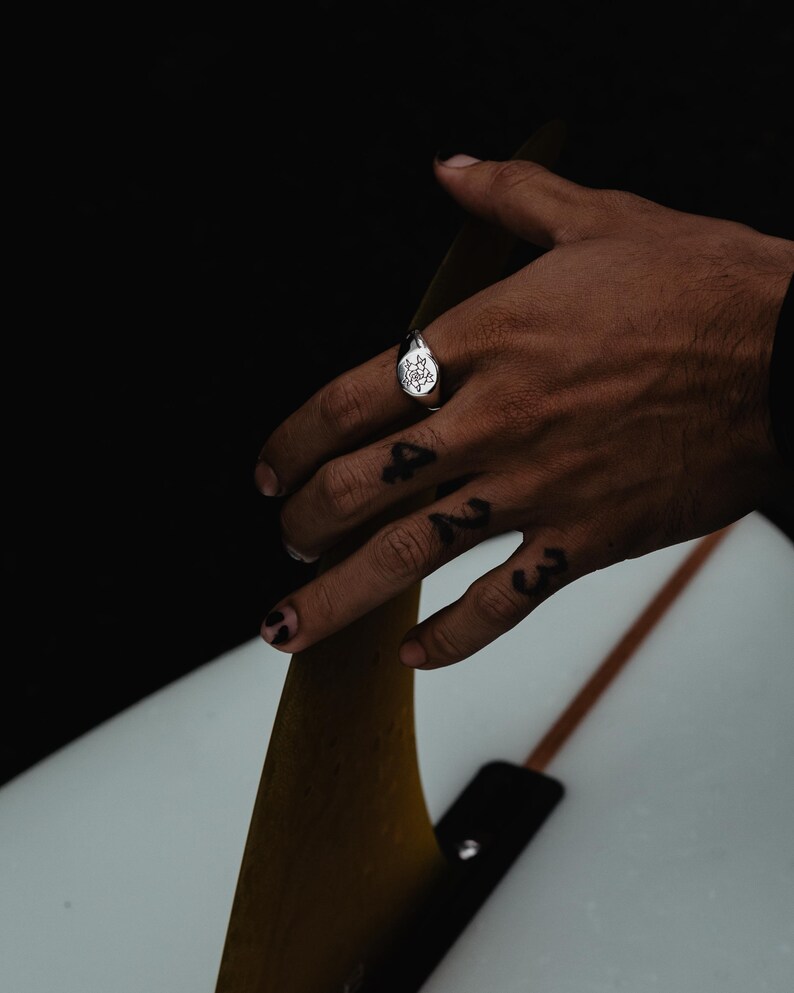 May include: A silver signet ring with a detailed design on the face. The ring is worn on the index finger of a hand resting on a surfboard fin.