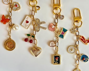 May include: Four gold-toned metal keychains featuring various charms including a letter M, a butterfly, a pearl, a sun, an ace of hearts card, a bow, cherries, a heart, a seashell, an envelope, a rose, a cowboy boot, and star motifs.