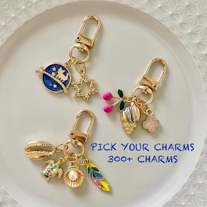 Può includere: Tre portachiavi dorati con ciondoli diversi. Il primo portachiavi ha un ciondolo blu e oro con un gatto e stelle, il secondo portachiavi ha un ciondolo rosa e verde con una conchiglia a righe e un fiore, e il terzo portachiavi ha un ciondolo a conchiglia, una tartaruga, una perla e una piuma. Il testo "PICK YOUR CHARMS 300+ CHARMS" è visibile nell'immagine.