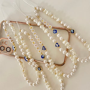 Può includere: Diverse catenine per telefono con perle con dettagli dorati e perline bianche con lettere che compongono nomi. Ogni catenina presenta un ciondolo con l'occhio blu. Una catenina dice "LOVE".