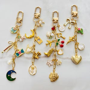 Puede incluir: Cuatro llaveros de metal dorado con varios dijes, incluyendo una luna creciente azul, un sobre rosa, una bota vaquera, una concha, un lazo blanco, una letra M, una cereza, un avión, un cactus y varias perlas pequeñas.