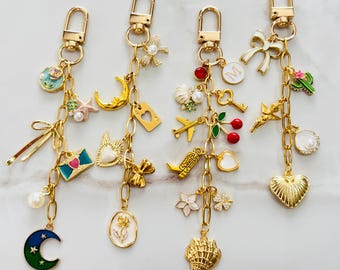May include: Four gold-tone metal keychains featuring various charms including a blue crescent moon, a pink envelope, a cowboy boot, a seashell, a white bow, a letter M, a cherry, an airplane, a cactus, and several small pearls.