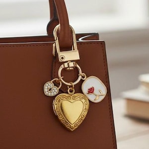 Può includere: Una borsa in pelle marrone con un ciondolo dorato. Il ciondolo presenta un medaglione a forma di cuore, un piccolo ciondolo a forma di cuore con strass e un ciondolo bianco con un disegno di rosa rossa. La borsa è appesa a una chiusura dorata.