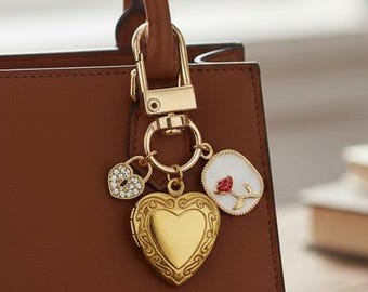 Könnte beinhalten: Eine braune Lederhandtasche mit einem goldfarbenen Anhänger. Der Anhänger hat ein herzförmiges Medaillon, einen kleinen herzförmigen Anhänger mit Strasssteinen und einen weißen Anhänger mit einem roten Rosenmuster. Die Tasche hängt an einem goldenen Verschluss.