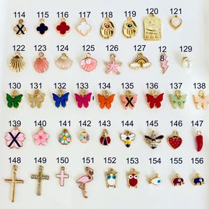 May include: A collection of gold-toned charms with various designs, including butterflies, flowers, crosses, and sea creatures. The charms are in different colors, such as pink, red, blue, and green. The image is taken from a top-down perspective.