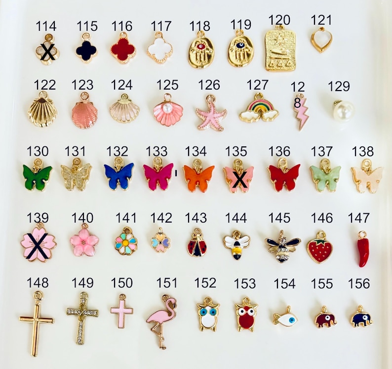 May include: A collection of gold-toned charms with various designs, including butterflies, flowers, crosses, and sea creatures. The charms are in different colors, such as pink, red, blue, and green. The image is taken from a top-down perspective.