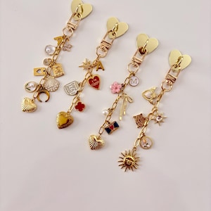 May include: Four gold-colored zipper pulls with heart-shaped tops and various charms. Charms include hearts, pearls, flowers, and a sun. The zipper pulls are on a white background.