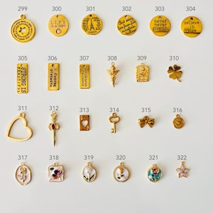 May include: A collection of gold-colored charms with various designs, including inspirational quotes, Eiffel Tower, a four-leaf clover, and floral motifs. Some charms have enamel details. The charms are numbered from 299 to 322.