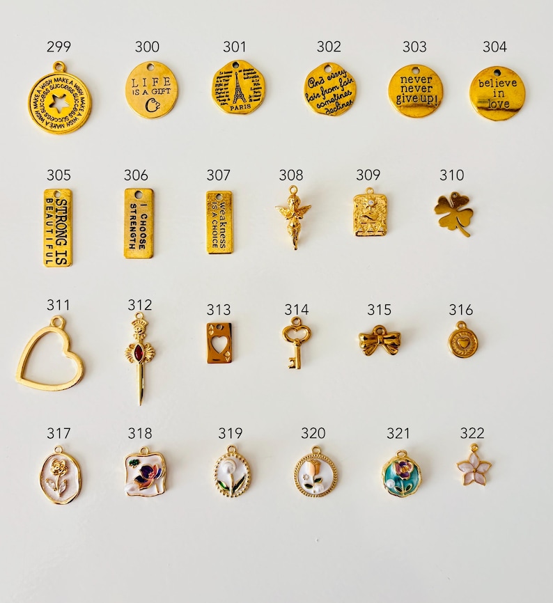 May include: A collection of gold-colored charms with various designs, including inspirational quotes, Eiffel Tower, a four-leaf clover, and floral motifs. Some charms have enamel details. The charms are numbered from 299 to 322.