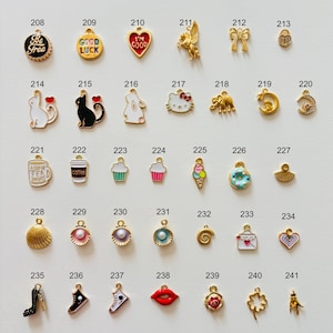 May include: A collection of gold-toned charms with enamel details. Designs include a heart, a cat, a cupcake, a high heel, a four-leaf clover, and a lips charm. Some charms have text like "Be Free" and "Good Luck".