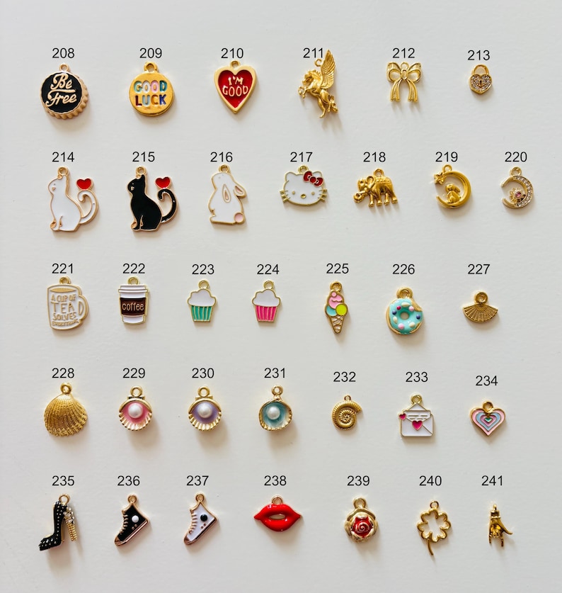 May include: A collection of gold-toned charms with enamel details. Designs include a heart, a cat, a cupcake, a high heel, a four-leaf clover, and a lips charm. Some charms have text like "Be Free" and "Good Luck".