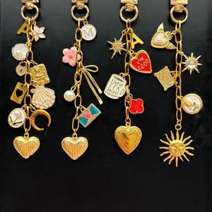 May include: Four gold-toned zipper pull charms with various dangling charms. Each charm features a heart-shaped pull. The dangling charms include letters, flowers, hearts, and other decorative elements. The charms are on a black background.