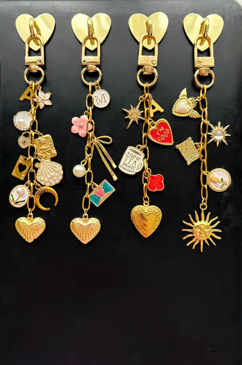 May include: Four gold-toned zipper pull charms with various dangling charms. Each charm features a heart-shaped pull. The dangling charms include letters, flowers, hearts, and other decorative elements. The charms are on a black background.