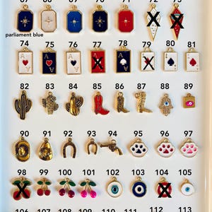 May include: A collection of gold-toned charms with various designs, including playing card suits, cacti, cowboy boots, and cherries. Some charms feature colorful enamel accents, such as blue, pink, and red. The charms are displayed on a white surface.