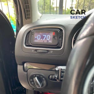 Op de afbeelding: Zwart dashboard van een auto met een digitale boostmeter die "-0,70 bar" weergeeft. De meter is in een ventilatieopening gemonteerd, met de tekst "Boost" eronder. De koplampbediening en een ventilatieopening zijn eronder zichtbaar. De tekst "CAR SKETCH" staat rechtsboven.