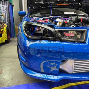 Puede incluir: Un Subaru Impreza WRX STI azul con un parachoques delantero negro, faros negros y un emblema "WRX" negro y rojo en la parrilla delantera. El capó está abierto, revelando el motor.