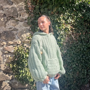Peut inclure: Un sweat à capuche vert clair en tricot épais avec une poche kangourou et une capuche à cordon. Le pull surdimensionné est fait d'une laine épaisse et a une coupe décontractée. Le sweat à capuche est porté avec un jean bleu clair.
