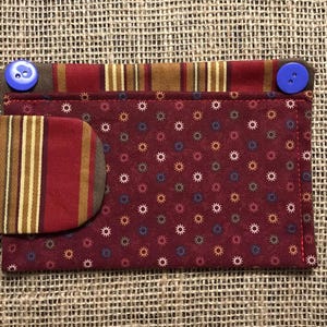 May include: A fabric wallet with a burgundy background and a pattern of small, colorful starbursts. The wallet has a striped pocket and is secured with two blue buttons.