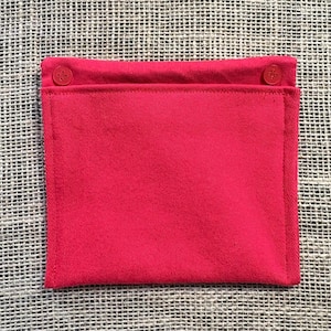 May include: A bright red, rectangular fabric pouch with a folded top secured by two matching buttons. The pouch is made of a solid, smooth material and is likely used for storage or carrying small items. The background is a textured, neutral-colored surface.
