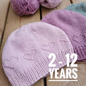 Könnte beinhalten: Gestrickte Mützen in verschiedenen Rosa- und Lilatönen mit Herzmustern. Das Bild zeigt auch Wollknäuel in passenden Farben. Auf der Mütze steht "2-12 YEARS".