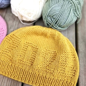 May include: A mustard yellow knit beanie featuring the word LOVE textured into the fabric using a combination of stockinette and reverse stockinette stitches. The hat has a ribbed brim and is positioned near balls of white, green, and blue yarn.