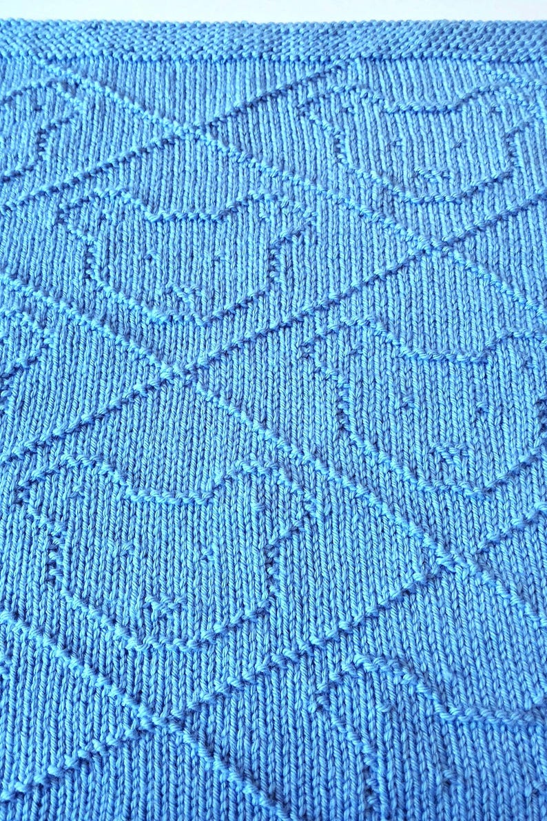 May include: Close-up of a knitted blue blanket with a geometric pattern. The blanket is a solid light blue color with a textured surface. The pattern consists of diagonal lines and shapes.