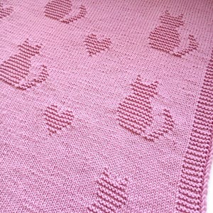 Può includere: Una coperta rosa lavorata a maglia con un motivo di gatti e cuori. I gatti sono stilizzati e ripetuti sulla coperta, con cuori intercalati. La coperta ha un bordo a coste.
