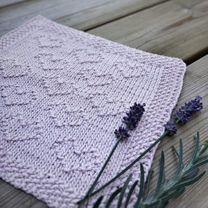 Pode incluir: Um pano de prato de malha rosa claro com um padrão texturizado, apoiado em uma superfície de madeira com ramos de lavanda.