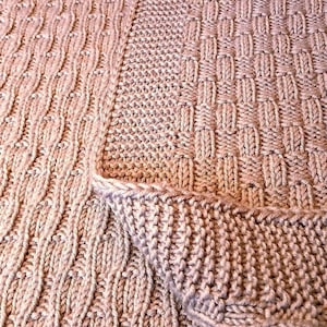 May include: Close-up of a knitted blanket in shades of beige and tan. The blanket displays a variety of textured patterns, including cable and basketweave designs. The folded edge shows the thickness and construction of the cosy, handmade item.
