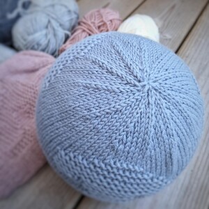 K&ouml;nnte beinhalten: Ein hellblaues, rundes Strickkissen mit strukturierter Oberfl&auml;che. Im Hintergrund befinden sich mehrere Wollkn&auml;uel in Grau-, Rosa- und Wei&szlig;t&ouml;nen. Das Kissen ist aus weichem Garn gefertigt und wahrscheinlich f&uuml;r die Inneneinrichtung gedacht.